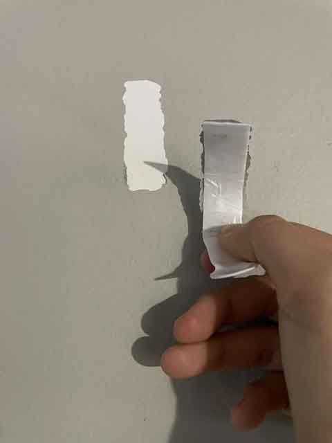 Hand peeling off a Command Strip from a wall, causing damage to the paint.
