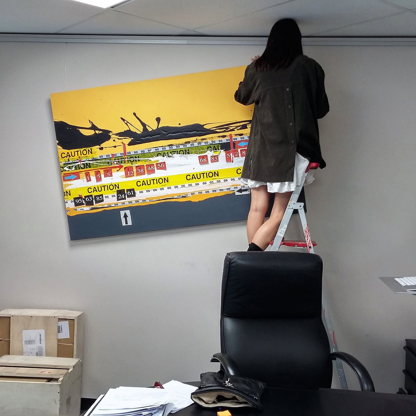 Woman on a ladder adjusting the height of a large yellow canvas in an office space using the flexible ArtiTeq sliding hook system