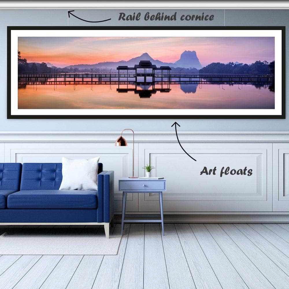 Long, framed landscape photo hanging on thin wires from an ArtiTeq Deco Rail picture rail concealed behind the cornice above a blue sofa.