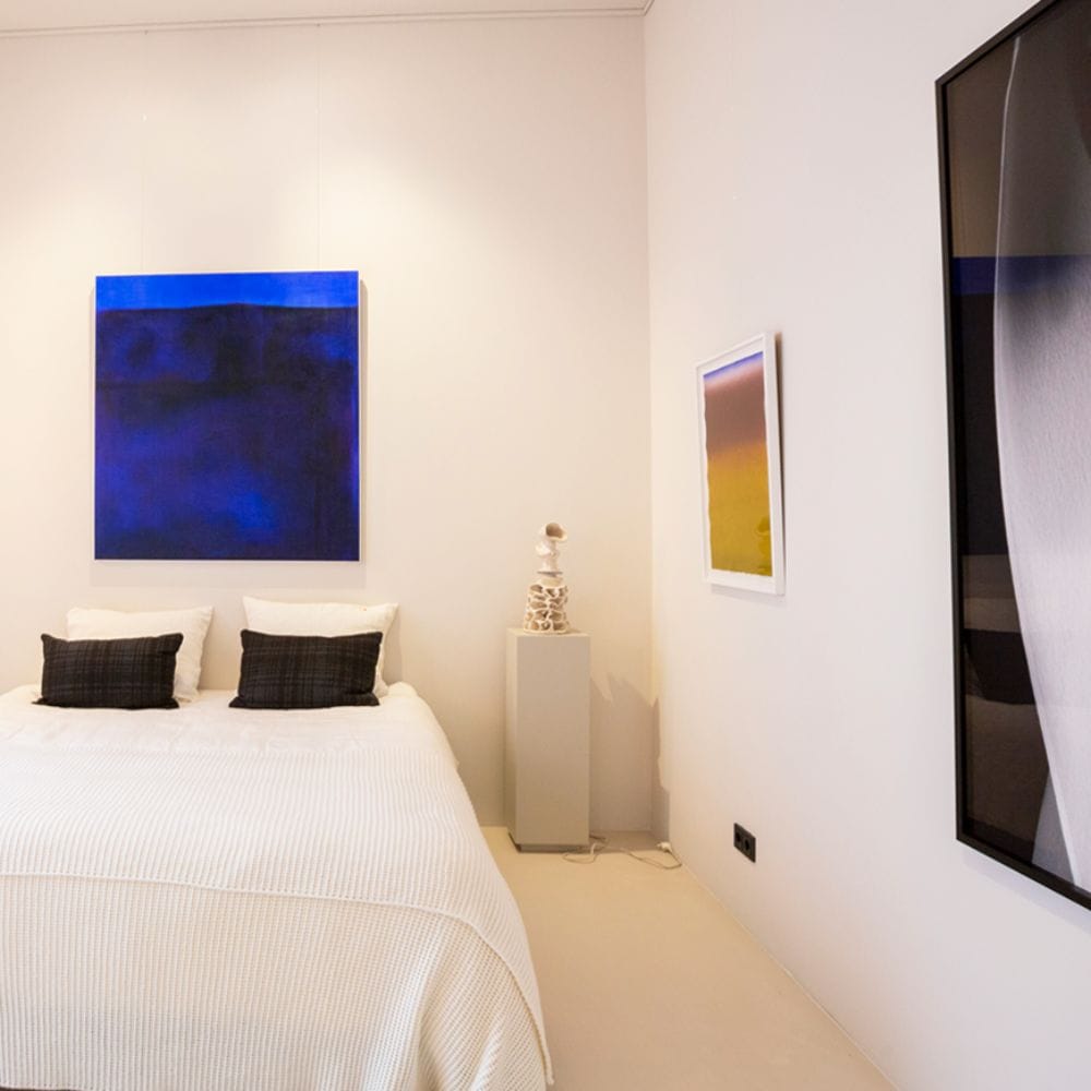 Bedroom with three artworks suspended from fine wires below picture rails, including a large blue piece above the bed.