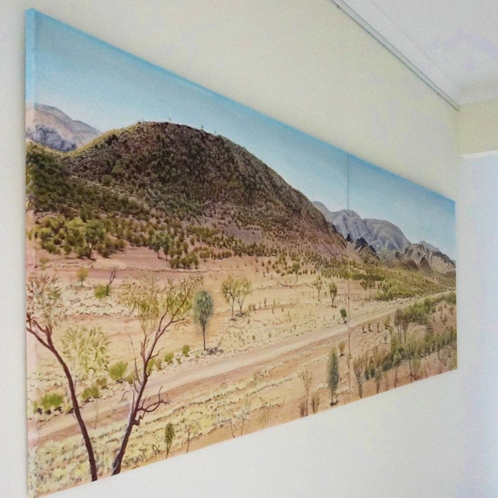 Large landscape artwork hanging on thin wires against a white hallway wall near the ceiling line.