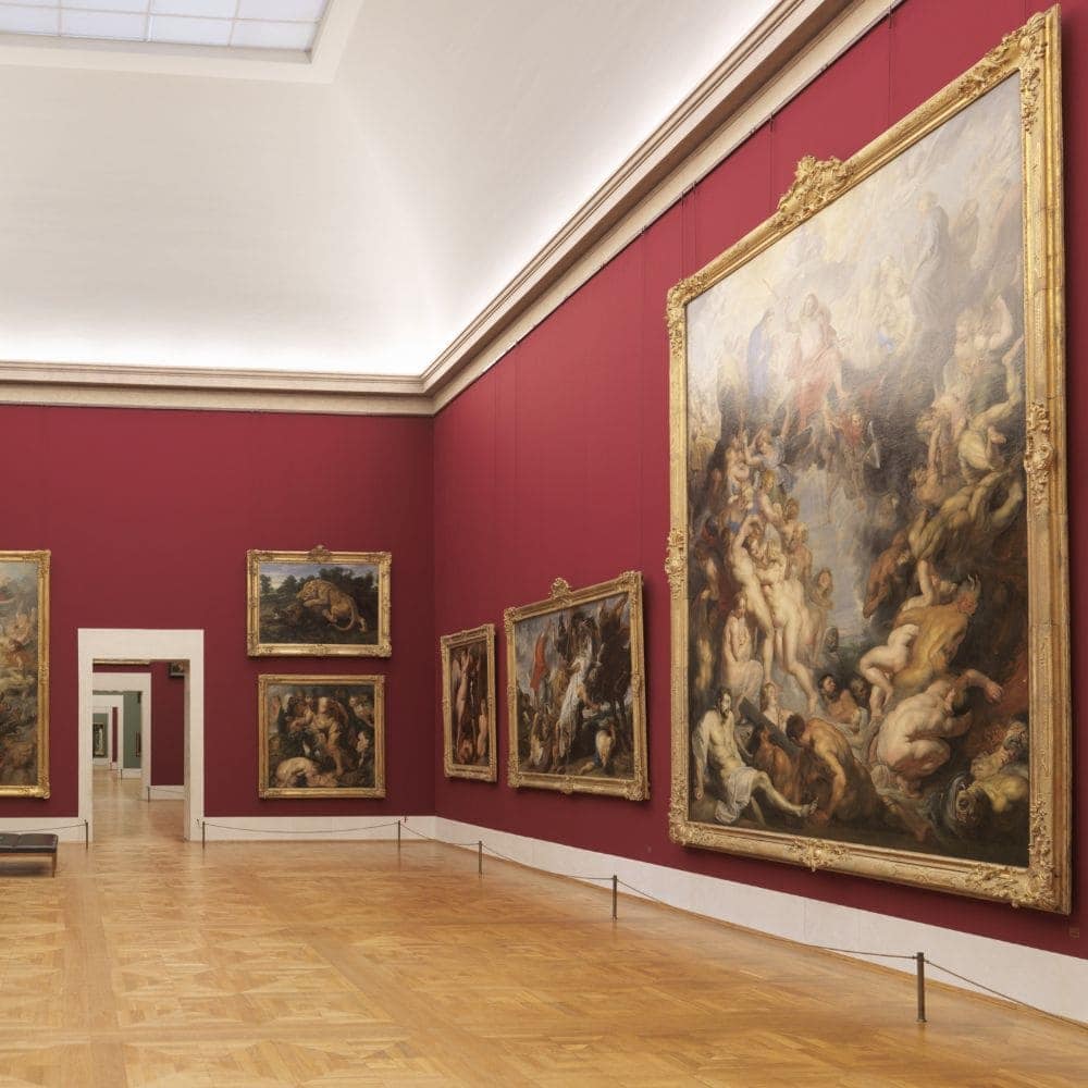 Large framed paintings hanging on wires from a slim rail in a museum gallery with deep red walls.